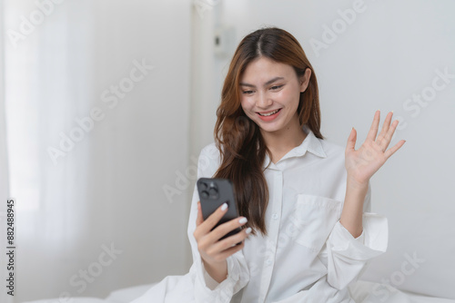 Young woman is sitting in bed having a video call on her smartphone and waving at the camera with a cheerful expression