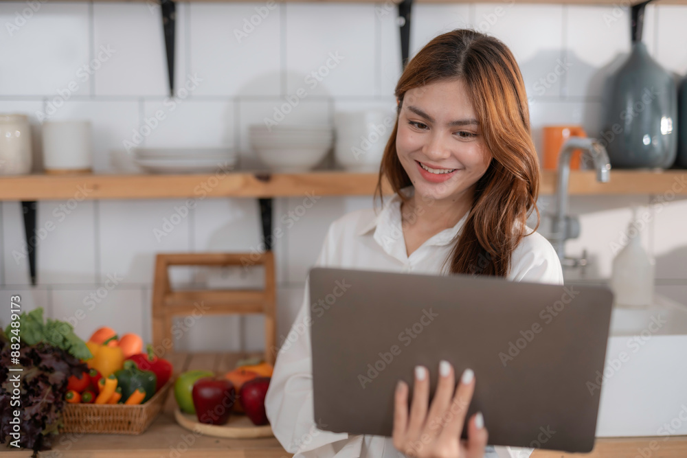 Young woman smiles in her cozy kitchen, holding a laptop while browsing for recipes online. Fresh produce decorates the counter, enhancing her culinary skills with modern technology Young woman smiles in her cozy kitchen, holding a laptop while browsing for recipes online. Fresh produce decorates the counter, enhancing her culinary skills with modern technology