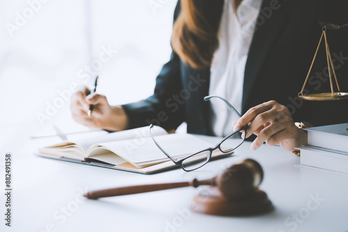 Lawyer is taking off her eyeglasses while writing in a notebook at her desk, with a gavel and scales of justice in the foreground