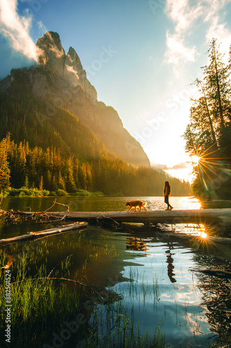 A Woman With A Dog Standing On The Log And looking at Majestic Mountain Peak of Mount Bering near Barclay Lake with Fog Haze on Sunny Day At Sunset photo