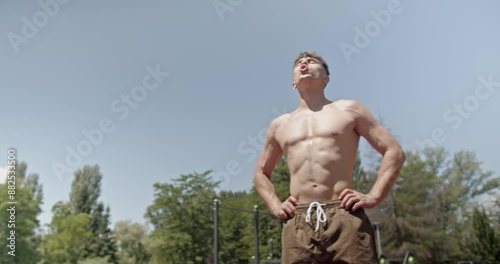A young man rests from an outdoor workout on a sunny day, standing shirtless and looking tired, showcasing his dedication to fitness and endurance.