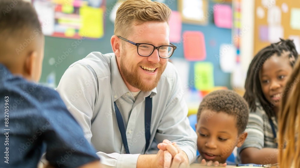 A bearded man with glasses and a lanyard smiles warmly as he engages with a group of children in a classroom setting. The children, of diverse backgrounds, are gathered around the man, listening inten A bearded man with glasses and a lanyard smiles warmly as he engages with a group of children in a classroom setting. The children, of diverse backgrounds, are gathered around the man, listening inten