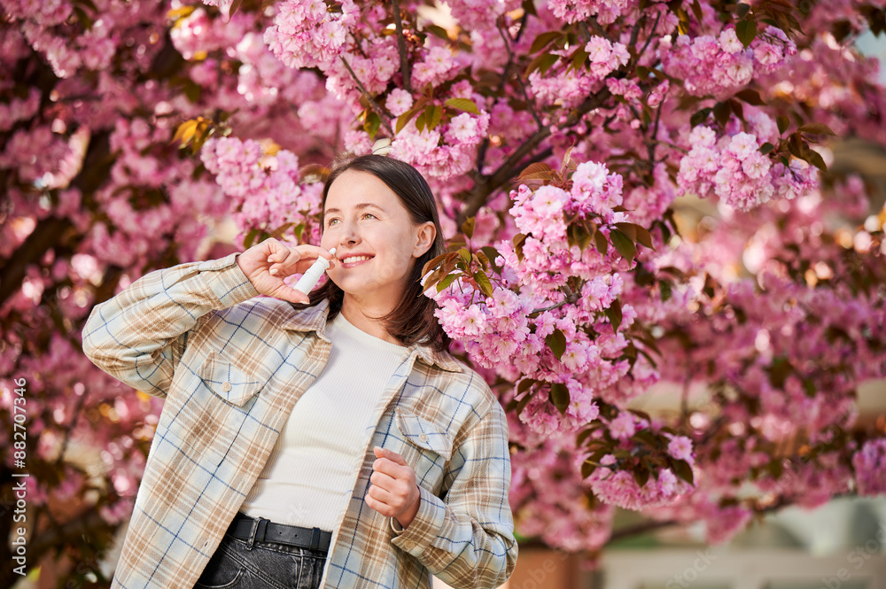 Woman allergic using medical nasal drops, suffering from seasonal allergy at spring in blossoming garden. Young woman treating runny nose in front of blooming tree outdoors. Spring allergy concept. Woman allergic using medical nasal drops, suffering from seasonal allergy at spring in blossoming garden. Young woman treating runny nose in front of blooming tree outdoors. Spring allergy concept.