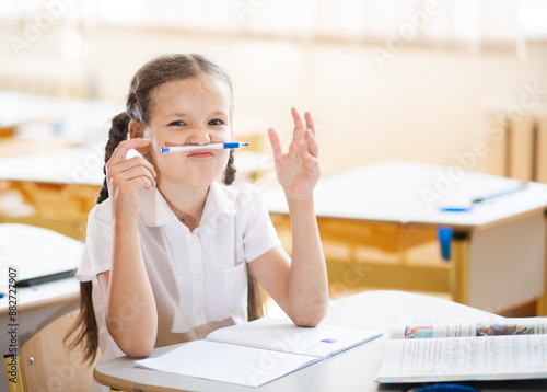 Back to school, a first-grader girl funnyly clasps her pen with her lips. A schoolgirl jokes in class. Comic school photo. School supplies on a child's desk.
 photo