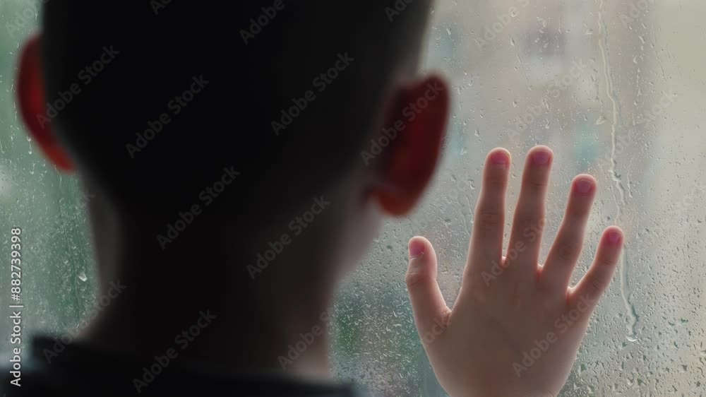 A bored boy leaned his palm against the window glass behind which it was raining heavily. autumn melancholy due to the rain outside the window A bored boy leaned his palm against the window glass behind which it was raining heavily. autumn melancholy due to the rain outside the window