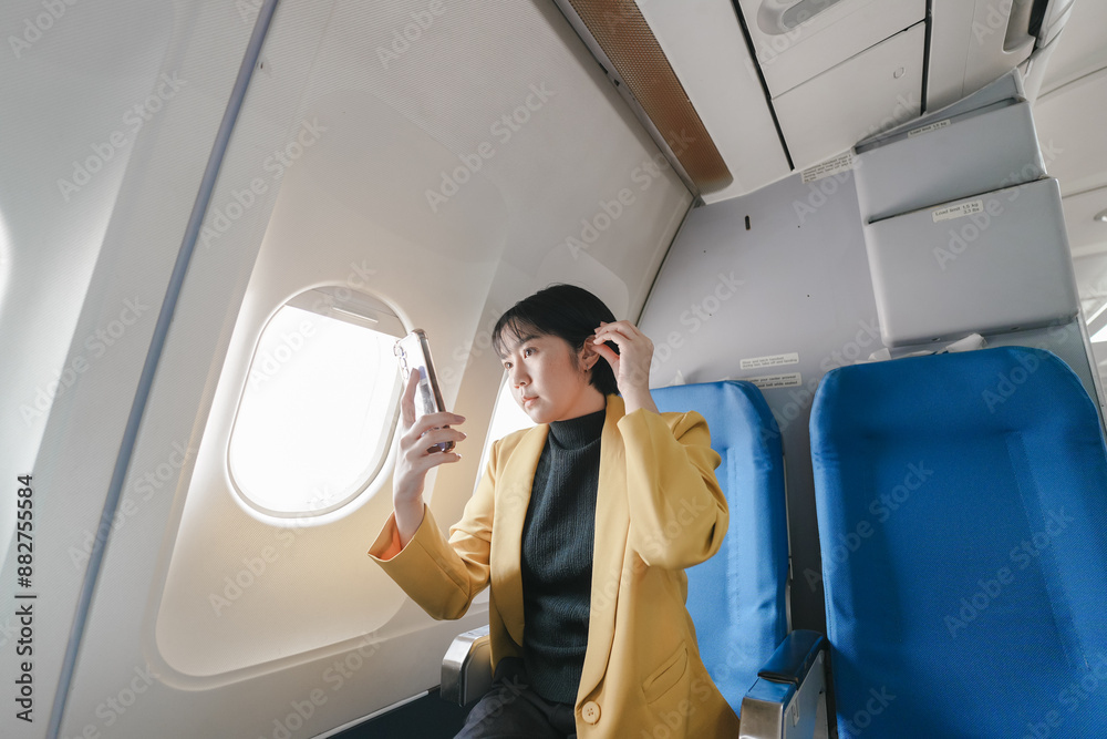 A woman is sitting in an airplane, looking at her phone. She is wearing a yellow jacket and black pants A woman is sitting in an airplane, looking at her phone. She is wearing a yellow jacket and black pants