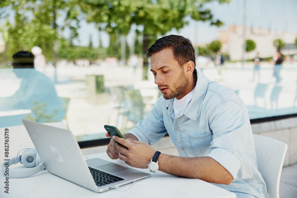 Fototapeta premium Young man working remotely on laptop while checking smartphone at outdoor cafe, focused and multitasking