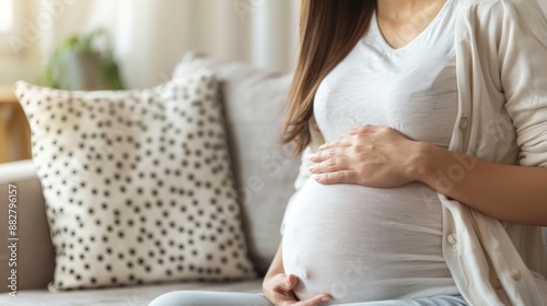 Close-up of a happy pregnant woman sitting on the sofa with a smile and holding her belly with both hands at home in the living room. copy space Free Download Close-up of a happy pregnant woman sitting on the sofa with a smile and holding her belly with both hands at home in the living room. copy space - GFXLoad