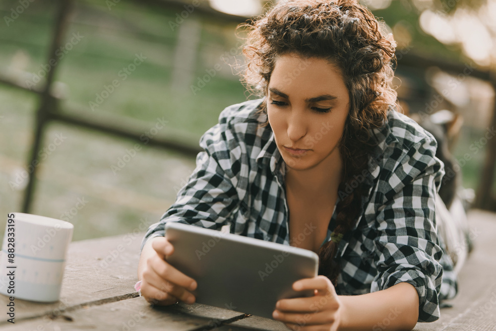 Woman with curly braided hair, wearing a checkered shirt, lies on her stomach, engrossed in a tablet. Her focused expression highlights her deep concentration, with a coffee mug beside her Woman with curly braided hair, wearing a checkered shirt, lies on her stomach, engrossed in a tablet. Her focused expression highlights her deep concentration, with a coffee mug beside her
