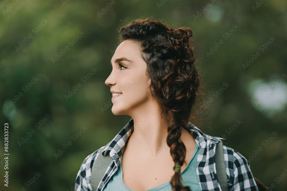 Woman with braided curly hair, wearing a checkered shirt and green top, smiles while observing her surroundings. Her backpack and relaxed demeanor indicate a pleasant outdoor adventure