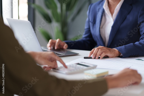 A professional setting featuring two individuals working at a table. They are engaged in reviewing documents and using a calculator. Laptops are open on the table.