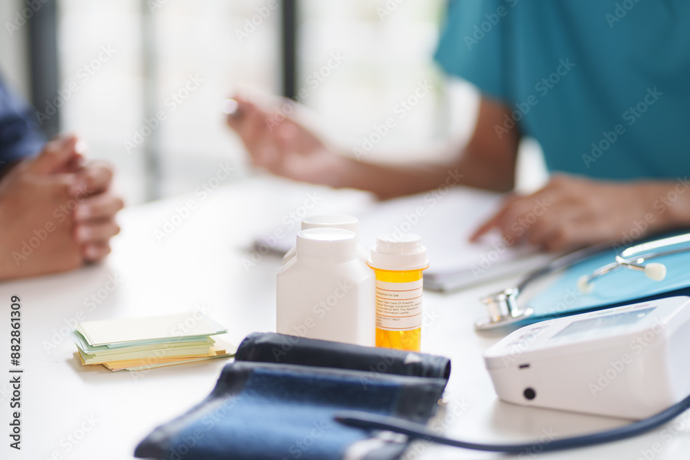 A doctor in a blue uniform discussing with a patient during a consultation. Medical bottles and documents are on the table. The patient’s hands are clasped together A doctor in a blue uniform discussing with a patient during a consultation. Medical bottles and documents are on the table. The patient’s hands are clasped together
