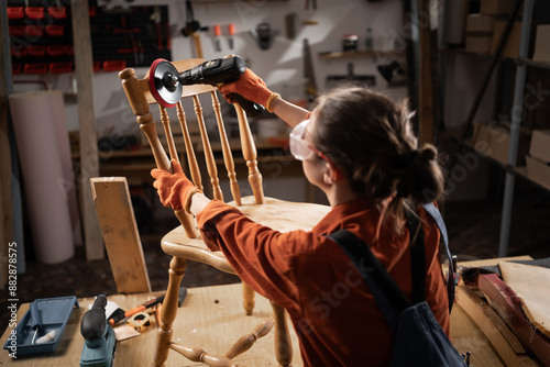 Woman using sander with a metal round bristle disk in a carpenter's workshop, sanding a wooden chair in the process of producing photo