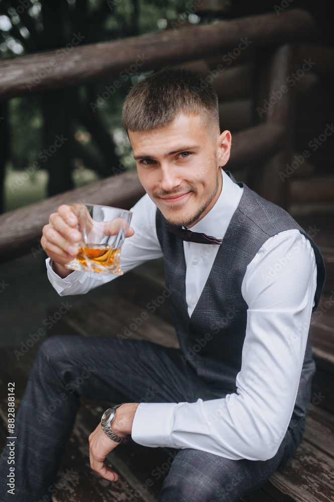 Wedding photography, portrait of a stylish smiling young groom, male businessman in a gray suit with a glass of whiskey. Wedding photography, portrait of a stylish smiling young groom, male businessman in a gray suit with a glass of whiskey.