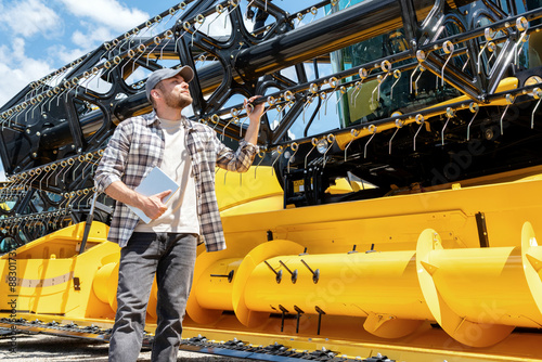 Caucasian adult man with digital tablet in hand looking at a yellow combine harvester at a farm machinery dealer. Ag equipment dealer or supplier.