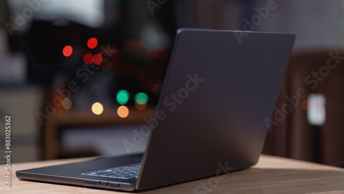 A laptop is open on a wooden table. The laptop is black and the screen is off. The table is in a dimly lit room