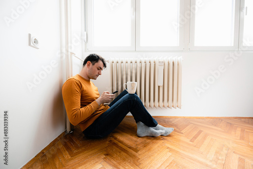 Man Sitting on Hardwood Floor Using Phone Near Radiator