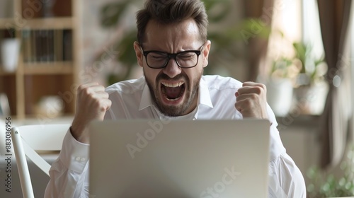 A man in a white shirt exhibits a strong emotional reaction, showing frustration and anger as he looks at his laptop screen, with clenched fists and an expression of aggravation.
