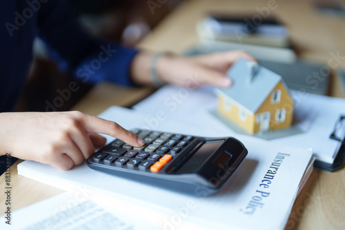 A house seller in a blue suit is working with a calculator at a desk. She is holding a piece of paper and appears focused on her calculations