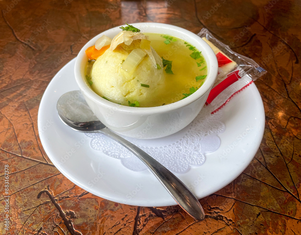 A bowl of matzo ball soup sits on a white plate with a spoon and a pack of saltine crackers. The soup is garnished with chopped vegetables and herbs, served in a clear broth on a textured metal table.
