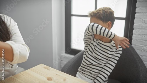 A woman offers a green apple to a reluctant boy in a striped shirt, capturing their interaction in a modern living room setting.