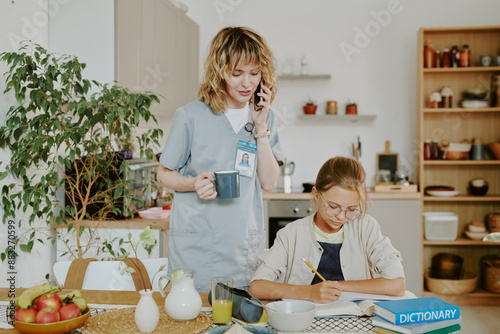 Doctor mother checking daughters homework and speaking on phone while daughter doing exercise in dining area photo