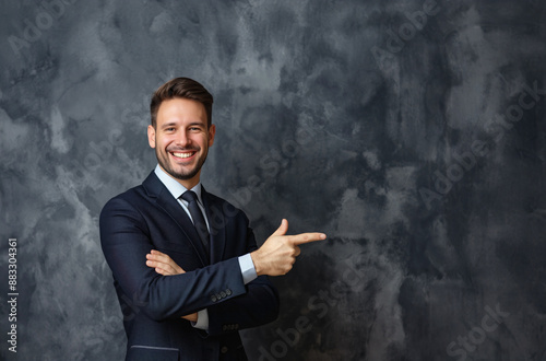 Smiling businessman pointing on dark textured background. Young businessman in a navy suit and tie, smiling and pointing to the side on a dark textured background