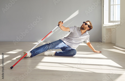 Disabled deaf visually impaired man falls down while walking with stick indoors. Vulnerable deafblind man in dark glasses with red white cane collapses on floor surface in room inside his apartment photo