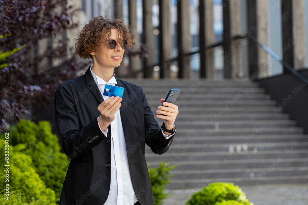Happy Caucasian man in stylish formal clothes using modern smartphone and credit card for online shopping. Smiling businessman guy with curly hair in sunglasses standing on street and making purchase. Happy Caucasian man in stylish formal clothes using modern smartphone and credit card for online shopping. Smiling businessman guy with curly hair in sunglasses standing on street and making purchase.