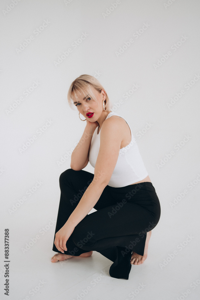  A blonde woman in a white crop top and black pants kneels on the floor, gazing to the side, set against a plain white background.