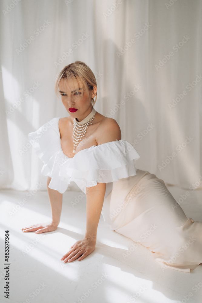 Elegant woman with blonde bob haircut and red lipstick posing on all fours in a white ruffled dress and pearl necklace, against a white background. Copy space... Elegant woman with blonde bob haircut and red lipstick posing on all fours in a white ruffled dress and pearl necklace, against a white background. Copy space...