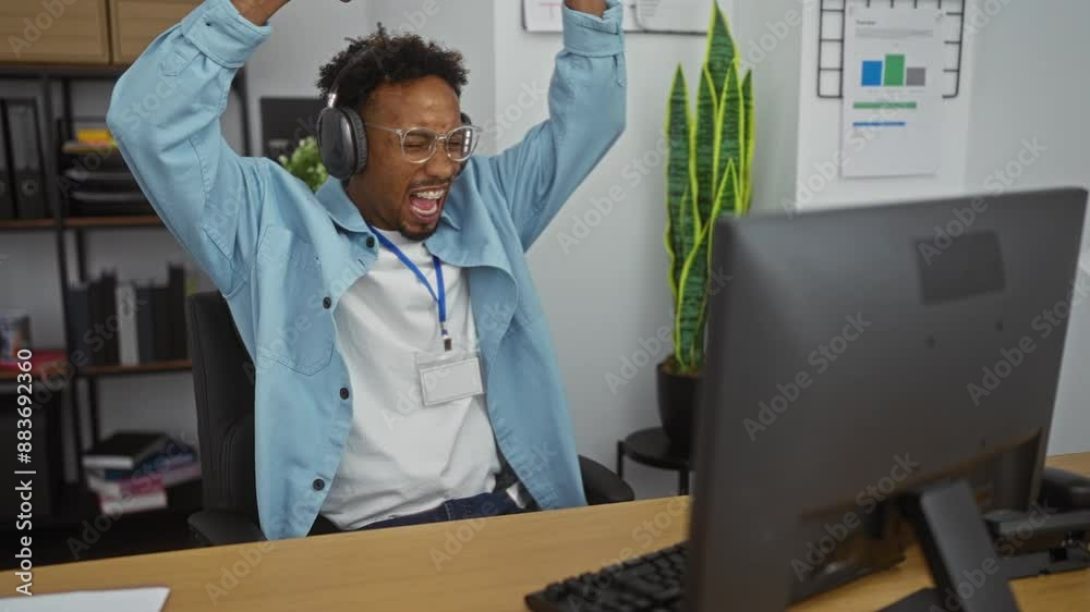 A young african american man with a beard and glasses sits at his desk in an office, wearing headphones and a blue shirt, looking focused as he engages with his computer. A young african american man with a beard and glasses sits at his desk in an office, wearing headphones and a blue shirt, looking focused as he engages with his computer.