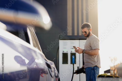 Adult male connects electric vehicle to charging station while checking his phone on a sunny day. photo