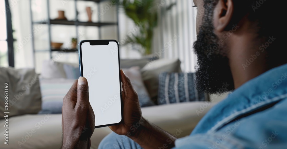 African American man using a smartphone in a cozy living room, with large windows providing natural light.