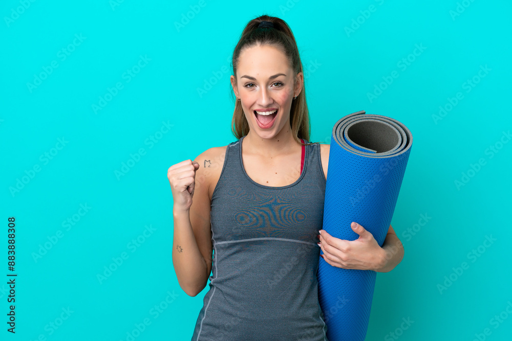 Young sport caucasian woman going to yoga classes while holding a mat isolated on blue background celebrating a victory in winner position Young sport caucasian woman going to yoga classes while holding a mat isolated on blue background celebrating a victory in winner position