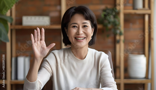 Beautiful Middle-Aged Asian Woman Sitting at Desk in Home Office Waving Hello on Video Call with Family or Friends, Smiling at Camera