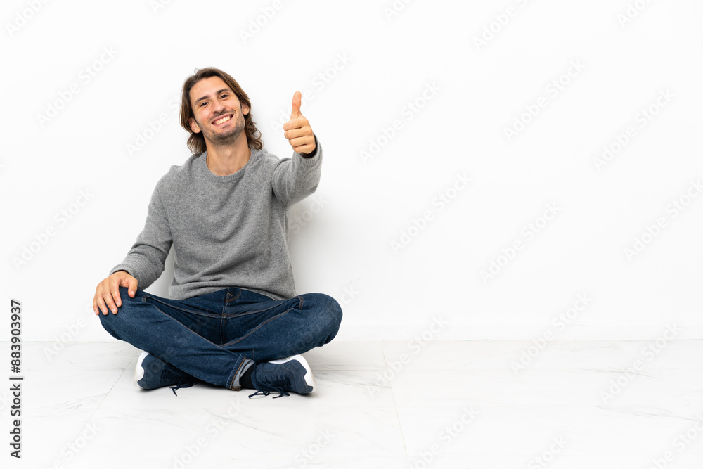 Young handsome man sitting on the floor over isolated background with thumbs up because something good has happened Young handsome man sitting on the floor over isolated background with thumbs up because something good has happened