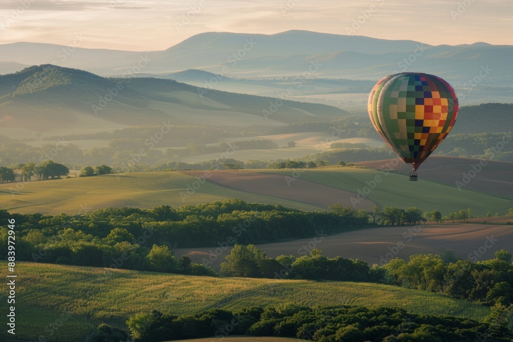 Fototapeta premium A hot air balloon drifting above rolling hills, with a softly blurred background of green fields and distant mountains. 