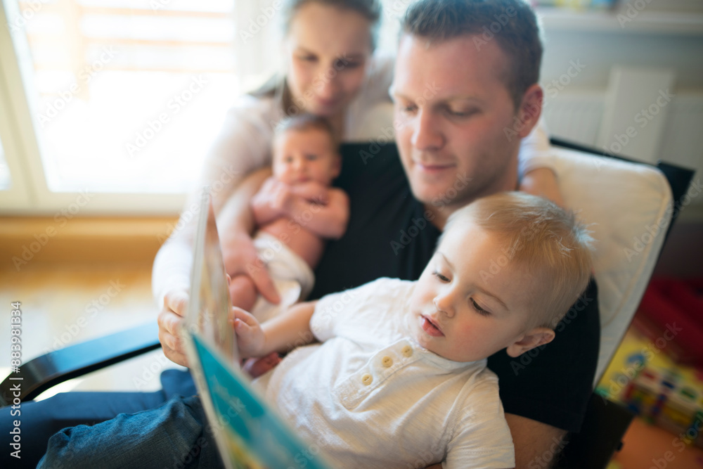 Obraz premium Nuclear family with neborn baby daughter reading book with older son. Mom and dad spending time with kids in room.