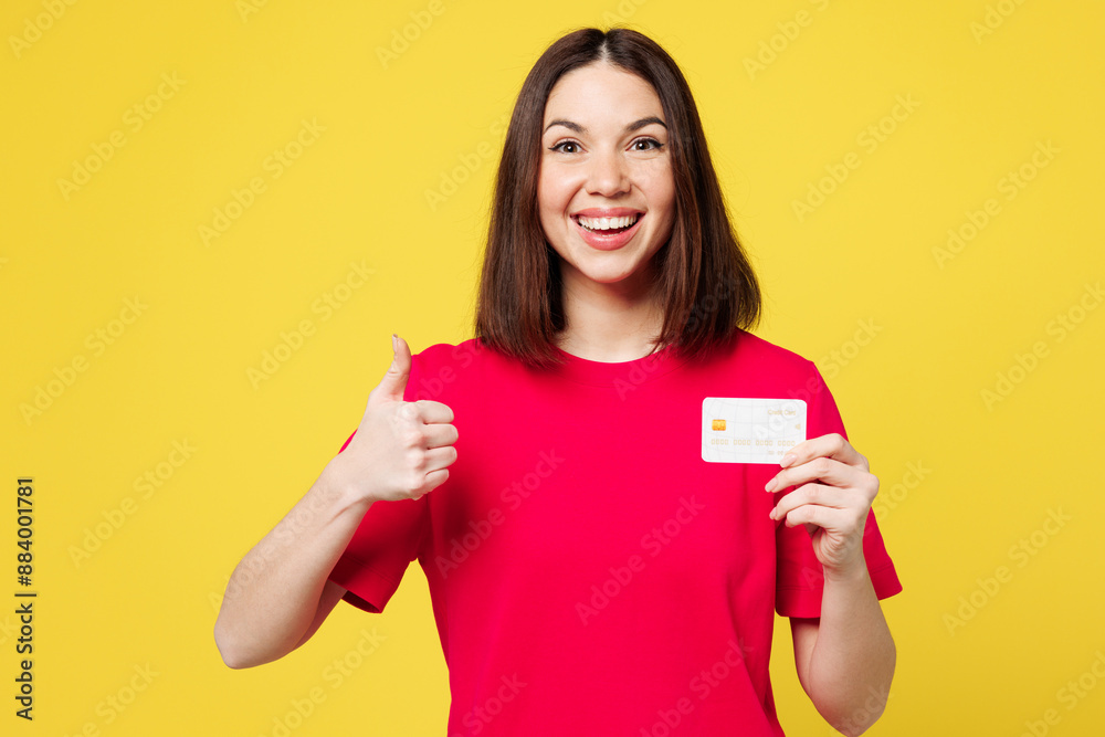 Young smiling fun happy woman she wear pink t-shirt casual clothes hold in hand mock up of credit bank card show thumb up isolated on plain yellow orange background studio portrait. Lifestyle concept. Young smiling fun happy woman she wear pink t-shirt casual clothes hold in hand mock up of credit bank card show thumb up isolated on plain yellow orange background studio portrait. Lifestyle concept.
