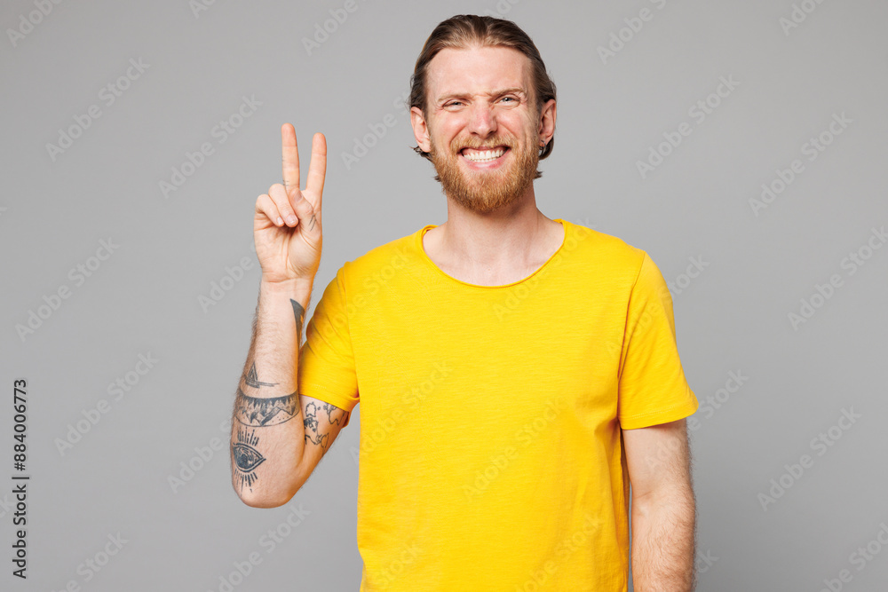 Young smiling happy fun joyful positive Caucasian man he wears yellow t-shirt casual clothes showing victory sign looking camera isolated on plain grey background studio portrait. Lifestyle concept. Young smiling happy fun joyful positive Caucasian man he wears yellow t-shirt casual clothes showing victory sign looking camera isolated on plain grey background studio portrait. Lifestyle concept.