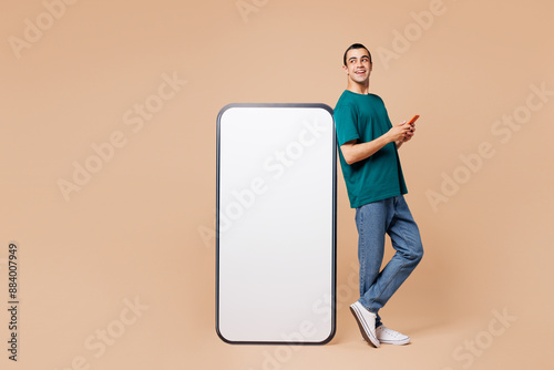 Full body side view fun young middle eastern man wear blue t-shirt casual clothes big huge blank screen mobile cell phone with workspace area use smartphone isolated on plain pastel beige background. photo