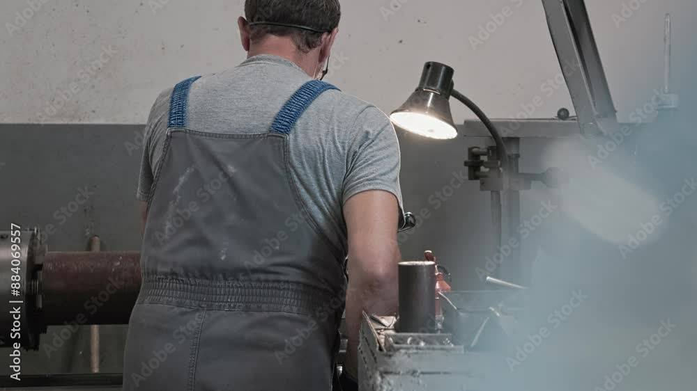 An individual is intently working on a lathe machine in a workshop, illuminated by a lamp, wearing overalls and safety goggles, showcasing concentration and craftsmanship. An individual is intently working on a lathe machine in a workshop, illuminated by a lamp, wearing overalls and safety goggles, showcasing concentration and craftsmanship.
