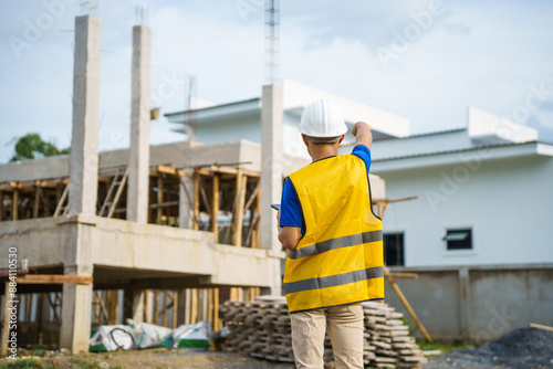 An architect, wearing a hardhat and safety vest, checks a laptop and house plan paper on a clipboard. Blueprints and construction documents lie nearby, emphasizing safety and precision in building.