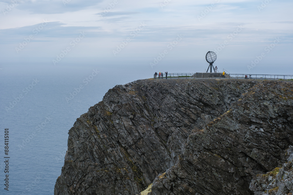 Fototapeta premium A group of tourists stand on the edge of a cliff, admiring the iconic globe monument at North Cape in Norway. The vast ocean stretches out before them, under a cloudy sky.