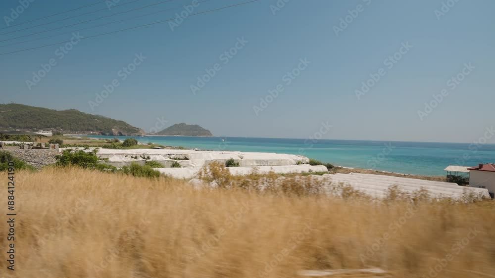 Multiple Greenhouses by the Sea, Viewed from a Moving Car. The Clear Blue Water and Distant Hills Create a Scenic Backdrop, Highlighting the Contrast Between Agriculture and Coastal Beauty. Multiple Greenhouses by the Sea, Viewed from a Moving Car. The Clear Blue Water and Distant Hills Create a Scenic Backdrop, Highlighting the Contrast Between Agriculture and Coastal Beauty.