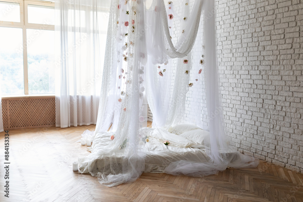 Bed with flowing curtains and garland flowers levitating, hanging flowers. Bedroom interior design.White Mattress for sleeping on the wooden floor with white linens in modern minimalist bright bedroom Bed with flowing curtains and garland flowers levitating, hanging flowers. Bedroom interior design.White Mattress for sleeping on the wooden floor with white linens in modern minimalist bright bedroom