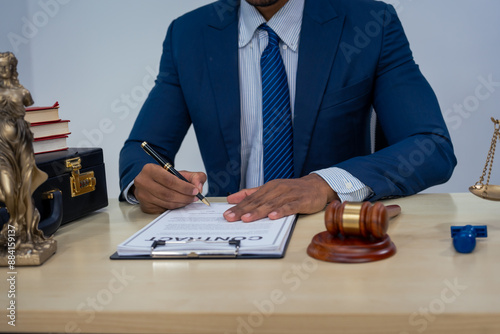 An Asian businessman in a formal suit works alone at a lawyer's desk, by golden scales, law books, leather bags, and a Lady Justice statue, emphasizing personal freedom and legal empowerment. photo