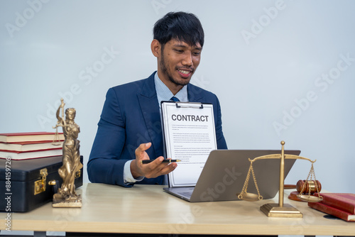 An Asian businessman in a formal suit works alone at a lawyer's desk, by golden scales, law books, leather bags, and a Lady Justice statue, emphasizing personal freedom and legal empowerment. photo