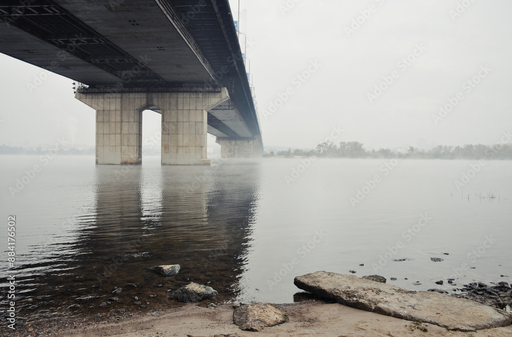 Bridge disappearing in a dense fog. Perspective view of the bridge from a bank of the river. Bridge over a river - low visibility on October morning. Bridge disappearing in a dense fog. Perspective view of the bridge from a bank of the river. Bridge over a river - low visibility on October morning.